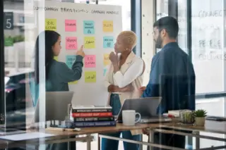 Diverse team in a modern coworking space discussing at a whiteboard with multilingual post-its, photographed candidly through glass with natural light.
