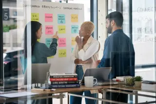 Diverse team in a modern coworking space discussing at a whiteboard with multilingual post-its, photographed candidly through glass with natural light.