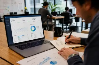 A professional working at a coworking desk, taking notes next to a laptop displaying KPI dashboards, in a naturally lit, active office environment.
