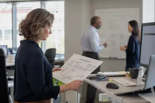 Professional woman reviewing an organizational chart at a standing desk, with two colleagues discussing by a whiteboard in a sunlit modern open office.