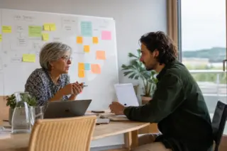 Two professionals engaged in a focused discussion by a window in a modern office, laptops open, whiteboard in the background, natural expressions and daylight creating an authentic work moment.