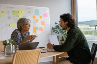 Two professionals engaged in a focused discussion by a window in a modern office, laptops open, whiteboard in the background, natural expressions and daylight creating an authentic work moment.