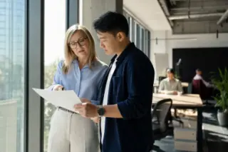 HR manager and project lead discussing a document by a window in a modern office, with a natural, candid atmosphere.