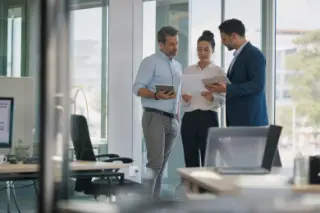 Three diverse managers discussing documents near a window in a modern office with natural Mediterranean daylight, captured candidly through a glass partition.