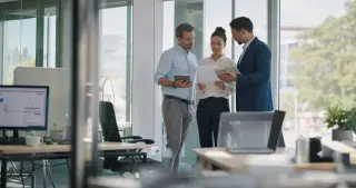 Three diverse managers discussing documents near a window in a modern office with natural Mediterranean daylight, captured candidly through a glass partition.