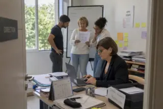 Open office scene with a woman reviewing applications at her desk, while three colleagues quietly discuss by a whiteboard, natural light and subtle Mediterranean details visible.