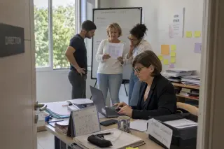 Open office scene with a woman reviewing applications at her desk, while three colleagues quietly discuss by a whiteboard, natural light and subtle Mediterranean details visible.