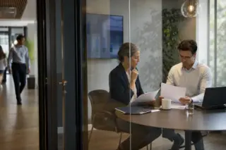 Two professionals reviewing documents separately in a glass-walled meeting room, offices and colleagues in soft focus background, natural daylight ambiance
