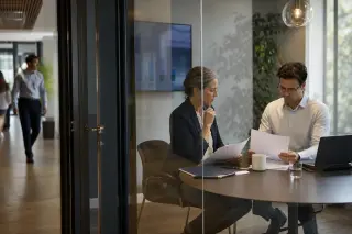 Two professionals reviewing documents separately in a glass-walled meeting room, offices and colleagues in soft focus background, natural daylight ambiance