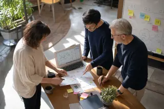Three professionals of different ages and backgrounds collaborating around a high table in a sunlit coworking space, reviewing project documents and a laptop, with a realistic, candid atmosphere.