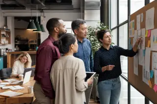 Four professionals in a coworking space discuss documents at a corkboard during an informal strategy session, daylight streaming through a large window.