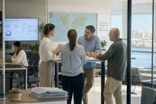 Professionals of different backgrounds in a modern open workspace engaged in a casual discussion by a standing table, with natural daylight and an authentic, unposed atmosphere.