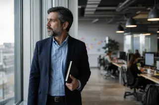Manager in business attire stands pensively by a window in a bright office while coworkers work in the background, natural daylight and subtle, realistic ambiance.