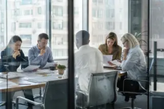 Plusieurs professionnels consultent chacun des documents RH internationaux dans un bureau, vus à travers une cloison vitrée.