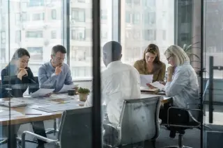 Plusieurs professionnels consultent chacun des documents RH internationaux dans un bureau, vus à travers une cloison vitrée.